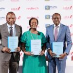 UBA, Redtech, MoMo PSB Expand Merchant Payment Access Across Nigeria Three adults in formal attire stand side by side, each holding a blue folder at a conference with sponsor logos in the background.