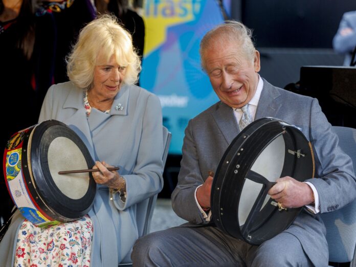 Two smiling seniors, a man in a gray suit and a woman in a pale blue coat, playing large frame drums together.