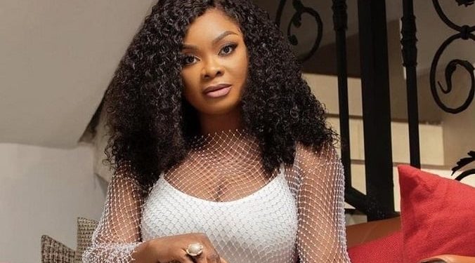 Portrait of a woman with voluminous curly hair wearing a white mesh top, seated on a sofa with red cushions in a living room setting, looking at the camera.