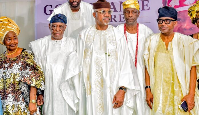 Five people in white and gold Nigerian agbada outfits posing at a cultural event; woman on left in a patterned dress and headwrap.