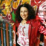 British Actress Emma Hussen Charged in Australia Over Alleged Meth Smuggling Operation Smiling woman with curly hair in a red jacket leaning against a graffiti-covered fence.
