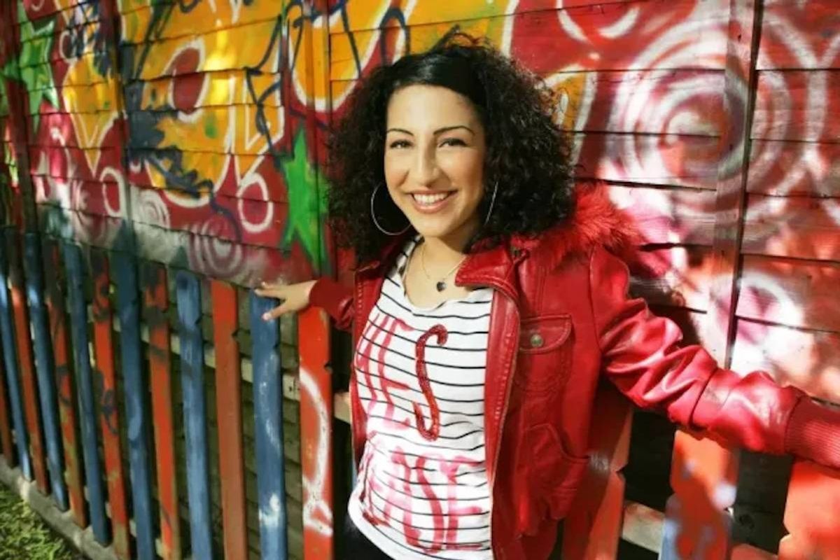 Smiling woman with curly hair in a red jacket leaning against a graffiti-covered fence.