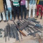 Police Bust Suspected Illegal Gun Factory Network In Akwa Ibom, Recover 15 Firearms Group of people standing behind a display of seized weapons laid out on the ground, including rifles, swords, and other metal items on a dusty street.