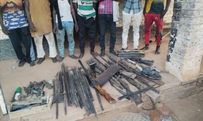 1001420175-400x240 Group of people standing behind a display of seized weapons laid out on the ground, including rifles, swords, and other metal items on a dusty street.