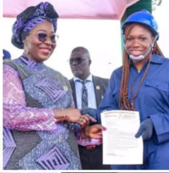 1001445243 Two women shake hands at an awards event: left in a purple patterned African dress and headwrap, right in a blue uniform holding a certificate; a suited man watches in the background.
