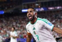 Kalajdzic’s Last-Gasp Equaliser Rescues Austria In Six-Goal World Cup Thriller Against Algeria Algerian national team player in a white kit celebrating a goal on the soccer field, arms outstretched and tongue out.