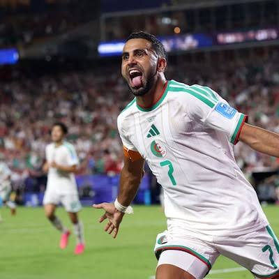 1002324045 Algerian national team player in a white kit celebrating a goal on the soccer field, arms outstretched and tongue out.