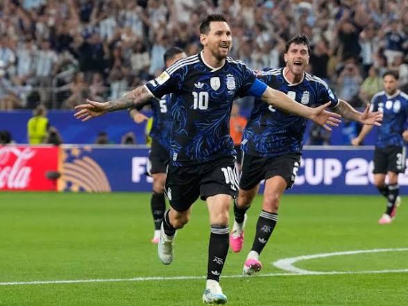 Two soccer players in blue jerseys celebrate a goal, arms outstretched as fans cheer in the stands.