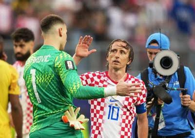 Croatian footballer in red-and-white checkered jersey (number 10) shakes hands with a green-clad goalkeeper on the pitch as a photographer films in the background.