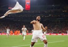 Morocco Knock Out Netherlands On Penalties To Set Up Canada Clash Soccer player in white kit celebrating on a green field with a flag flying in the air as fans cheer in the stadium