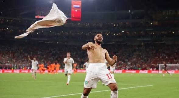 Soccer player in white kit celebrating on a green field with a flag flying in the air as fans cheer in the stadium