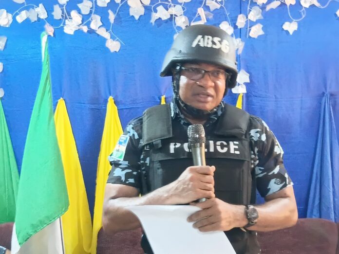 Police officer in camouflage uniform and helmet holding a microphone with a paper in front of a blue backdrop and yellow/green flags behind him, indoors.