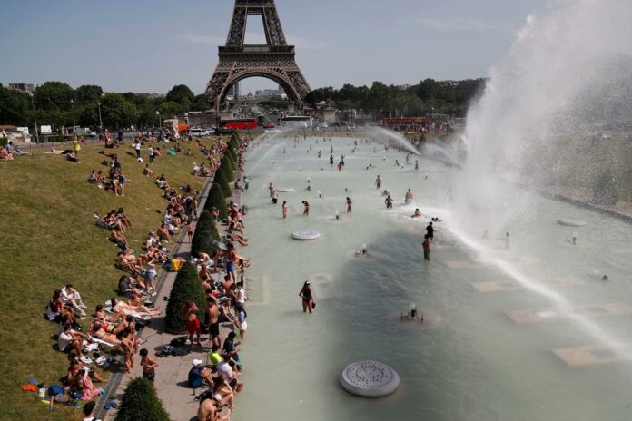 20260625_121121 Crowded Paris riverside near the Eiffel Tower, with people sunbathing on the grass and a large water fountain spraying in the foreground.