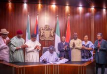 Tinubu Signs NIMC Act 2026 Into Law Group of dignitaries standing and clapping at a formal signing ceremony with national flags and crest in the background.