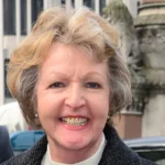 British Actress Dame Penelope Keith Dies After Cancer Battle Smiling older woman with short blond hair in a dark coat, standing outdoors with a stone building in the background.
