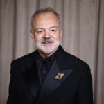 Irish Broadcaster Graham Norton Wins Court Order to Identify Facebook Account Behind False Posts Older man in a black tuxedo and bow tie standing against a brown curtain backdrop, wearing a gold decorative lapel pin with a green center, smiling slightly.