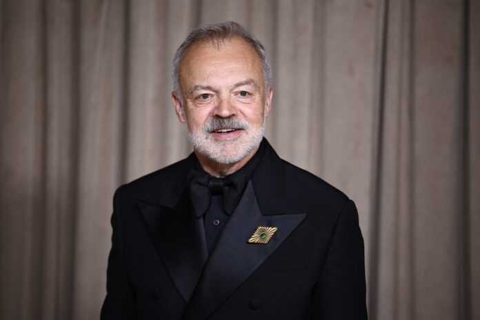 2238622520 Older man in a black tuxedo and bow tie standing against a brown curtain backdrop, wearing a gold decorative lapel pin with a green center, smiling slightly.