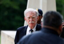 Trump Ex-Security Adviser John Bolton Pleads Guilty To Mishandling Classified Information Elderly man with white hair and mustache in a dark suit and striped tie, outdoors with a blurred figure nearby.