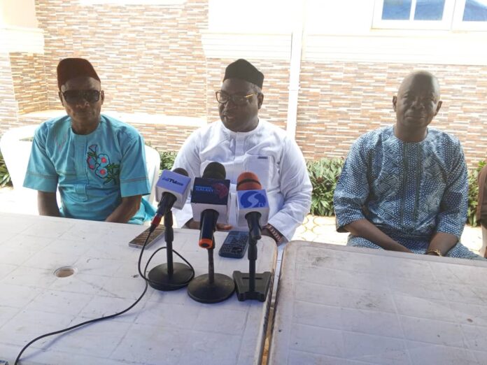 Three men sit at a white table outdoors with multiple microphones for a press conference.
