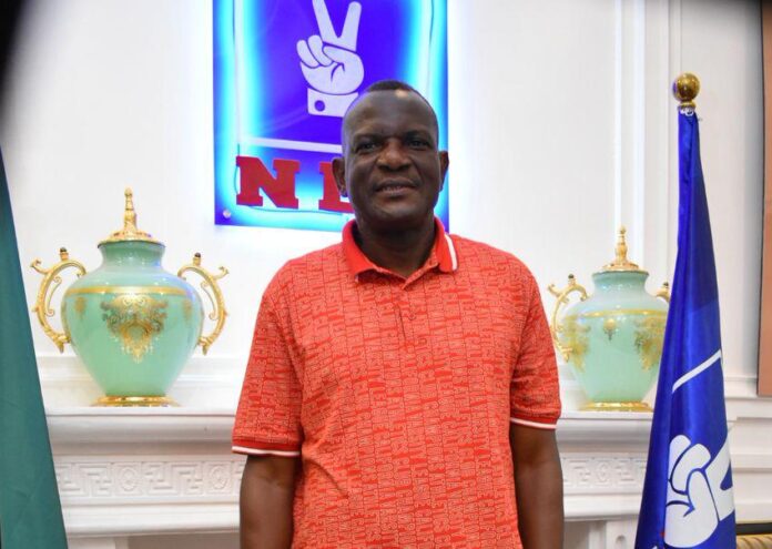 469975 Man in a red polo shirt posing in front of a blue illuminated sign, flanked by decorative green-and-gold vases and flags in a formal room.