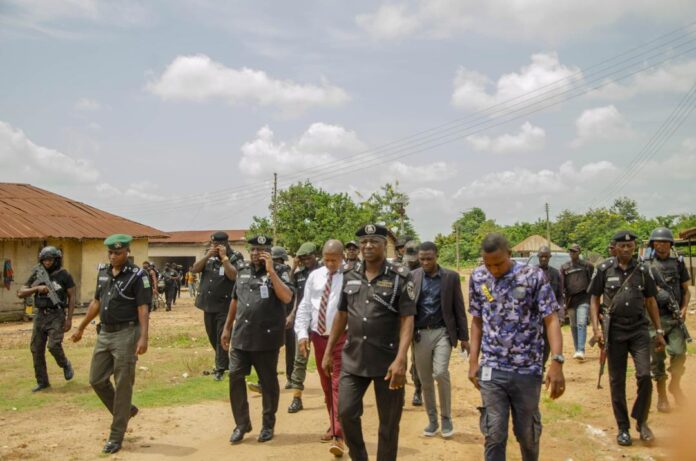 879227f6-cbc5-45d5-ad63-b90ca4177c76 Group of uniformed police officers walking down a dirt road in a rural area, flanked by a few civilians and onlookers.