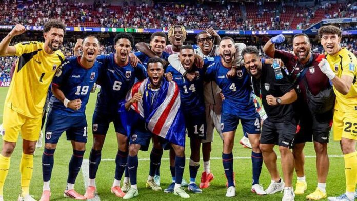 Soccer team in blue celebrating together on the field after a win, arms around each other in a stadium.