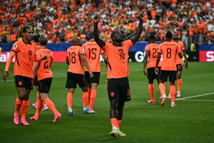 Soccer players in orange jerseys celebrate on the field with one player raising both arms as teammates applaud nearby. Crowd in orange in the stands behind them.