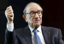 Former U.S. Federal Reserve Chair Alan Greenspan Dies at 100 Older man in a dark checked suit with glasses raises his right fist in a determined gesture against a dark background.