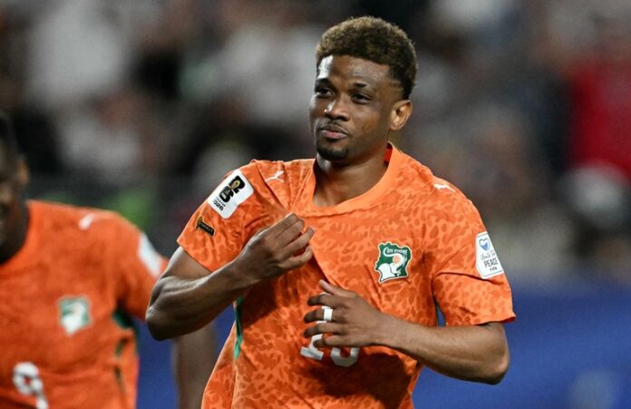 Soccer player in an orange patterned jersey adjusts his shirt on the field as a crowd watches in the background.
