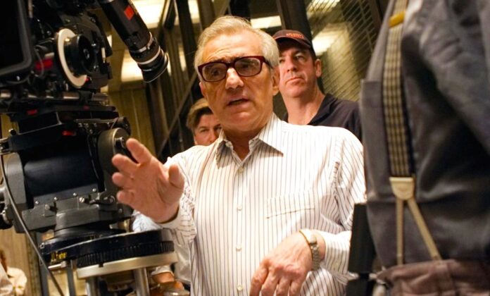 BELOVED-FILMMAKER_MARTIN-SCORSESE_BACKING_AI_COMPANY_ Man in striped shirt and glasses gestures toward a film camera on a movie set, with crew in the background.