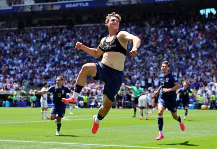 Athlete in mid-air celebrating a goal on a sunny soccer field with a cheering crowd in the stands behind him.”] ,