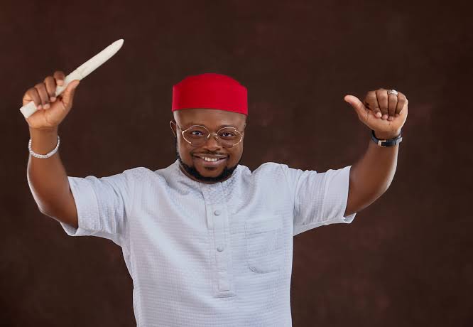 Chief (Dr) Ifeanyichukwu Odii Man in a red fez and white traditional outfit, smiling, holds a knife aloft in one hand and a thumbs-up in the other.