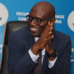 SEC Grants Approval-in-Principle to Seven Crypto Firms Under Regulatory Sandbox Smiling man in a navy suit and glasses sits with hands clasped, blue backdrop with Nigerian seal.
