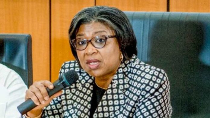 DMO-Hikes-Rates-on-Bonds-to-Meet-N1.2trn-Borrowing-Target Woman in a patterned blazer speaks into a handheld microphone at a panel discussion, wearing glasses and gold earrings.