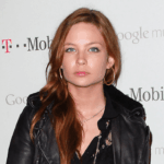 Lilo & Stitch Actress Daveigh Chase’s Cause Of Death Confirmed As AIDS Young woman with long red hair and hoop earrings, wearing a black leather jacket, posing in front of a backdrop with Google Music and T-Mobile logos.