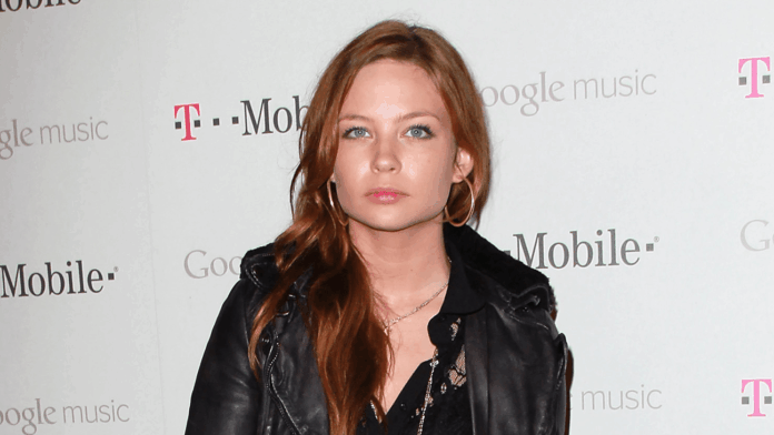 Young woman with long red hair and hoop earrings, wearing a black leather jacket, posing in front of a backdrop with Google Music and T-Mobile logos.