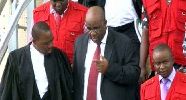 Emmanuel-Okoyomon Group of people on a staircase; a man in a suit with a red tie points as others in red vests and robes stand nearby.