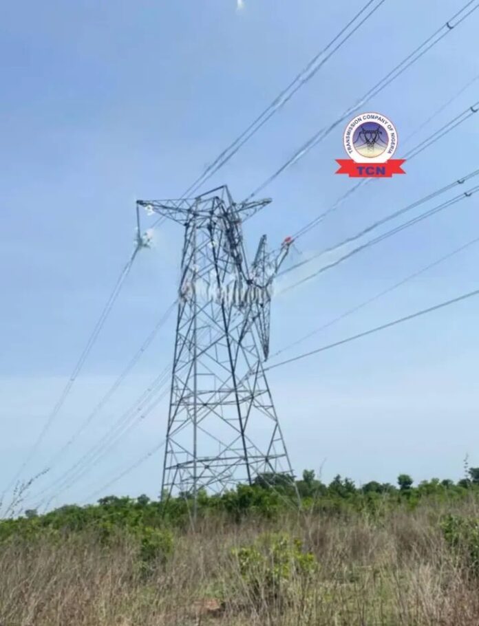 Tall metal transmission tower with high-voltage lines stretching into a rural field under a clear blue sky.