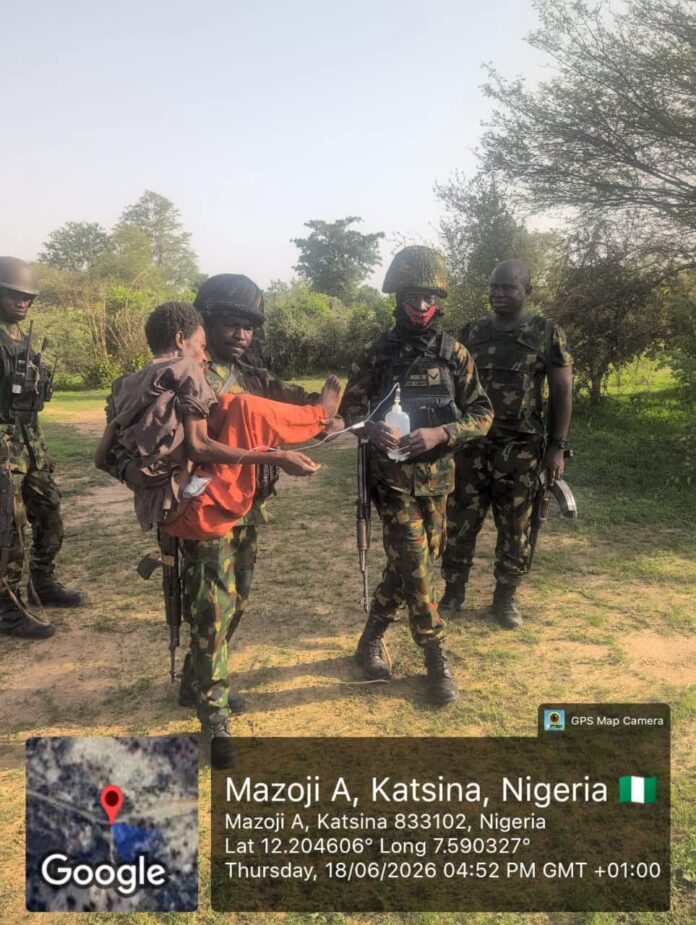 FB_IMG_1781858900846 Soldiers in camouflage with a man carrying a child wrapped in orange blanket on a grassy field; one soldier holds a water bottle while others watch.