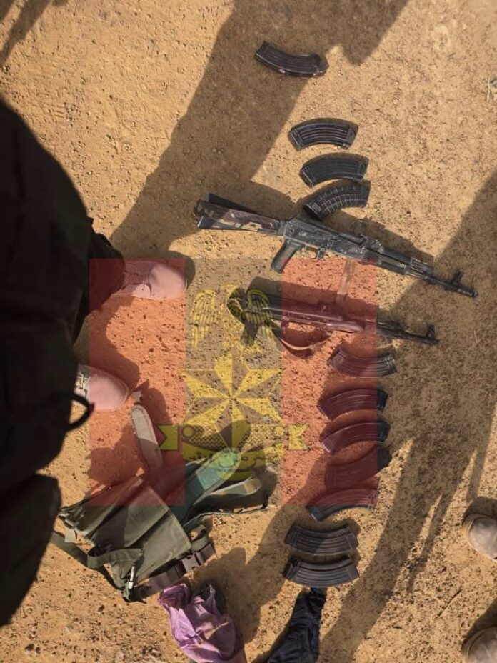 Assorted firearms magazines and an assault rifle laid out on dusty ground with backpacks nearby.