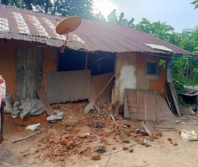 Rundown mud-walled house with a bent satellite dish on the corrugated metal roof, front yard full of debris and broken planks.