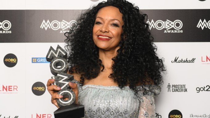 Smiling woman with curly black hair in a silver off-shoulder dress holds a MOBO Awards trophy on a branded backdrop.