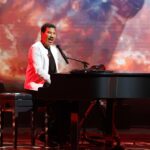 Lionel Richie Ends Minnesota Opening Night Show After Falling Ill Male performer in a white jacket plays a grand piano on a lit stage with a fiery, red-orange background behind him.