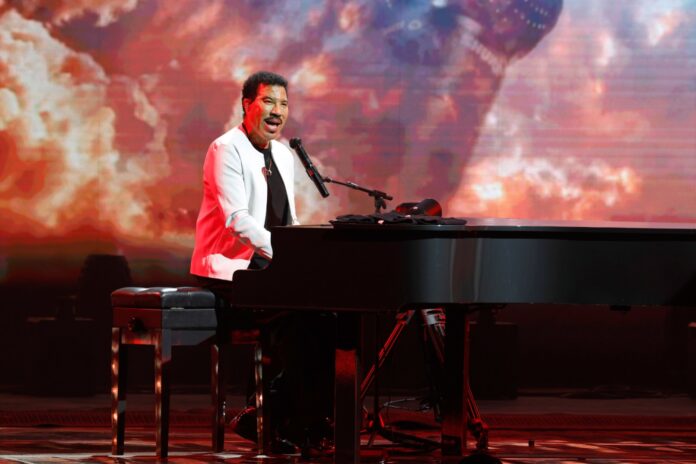 GettyImages-2276046632-1 Male performer in a white jacket plays a grand piano on a lit stage with a fiery, red-orange background behind him.