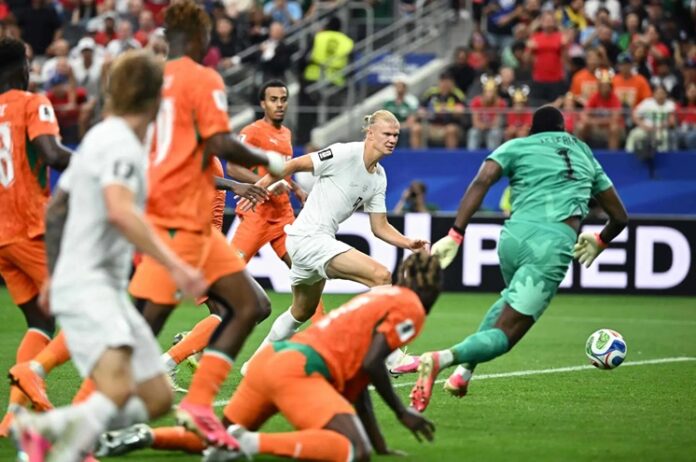 White-team attacker shoots toward goal as orange defenders converge and the teal goalkeeper guards the net.