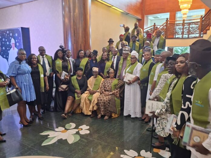IMG-20260601-WA0001 Group of people in green sashes posing for a formal group photo in a lobby with stairs and a chandelier behind them. They are dressed in varied formal attire, some holding books or programs, with two individuals seated in front.