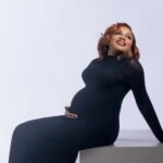Media Personality Ebuka Obi-Uchendu, Wife Announce Third Pregnancy Pregnant woman in a black form-fitting dress sits on a block, resting one hand on her baby bump and smiling toward the distance.