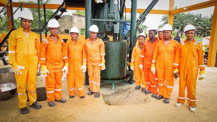 IMG-20260628-WA0094 Group of workers in orange coveralls and white hard hats standing around a large drilling machine outdoors.