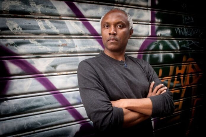 Man with arms crossed in a dark long-sleeve shirt poses in front of a graffiti-covered metal shutter with purple and white spray paint behind him.