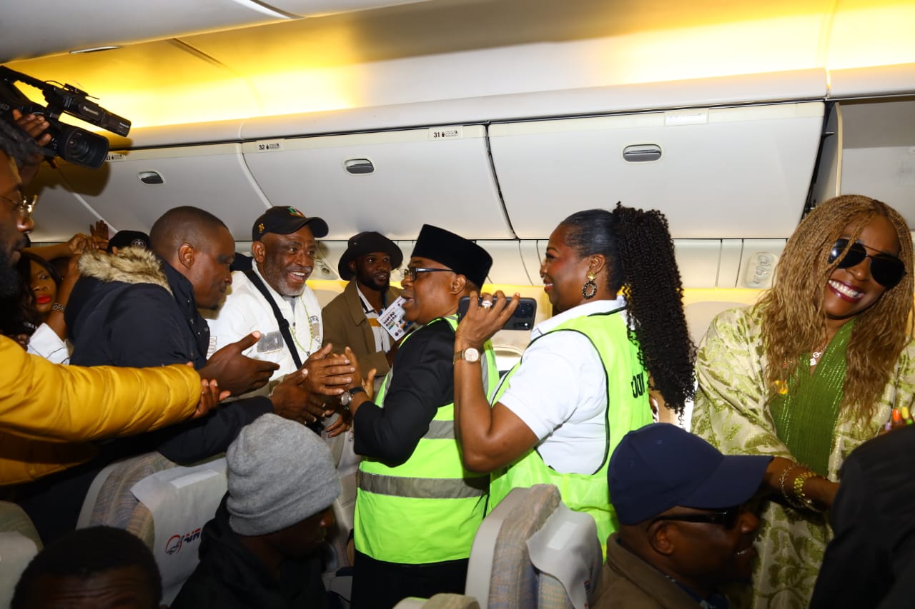 IMG_6015 - THEWILL NEWS MEDIA Group of passengers and crew on an airplane aisle exchanging handshakes and smiles during an arrival moment in the cabin, with a camera visible on the left.